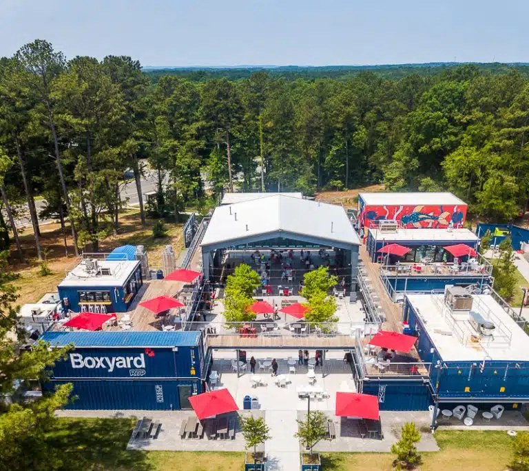 Aerial view of Boxyard RTP: blue shipping containers stacked two high around a courtyard of red umbrellas, under a covered pavilion, surrounded by pine woods